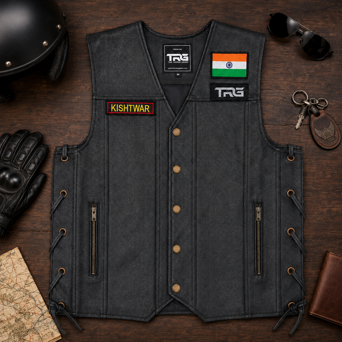 Premium flat lay of TRG black riding vest with Kishtwar embroidery patch, riding helmet, gloves, sunglasses, keys and map.