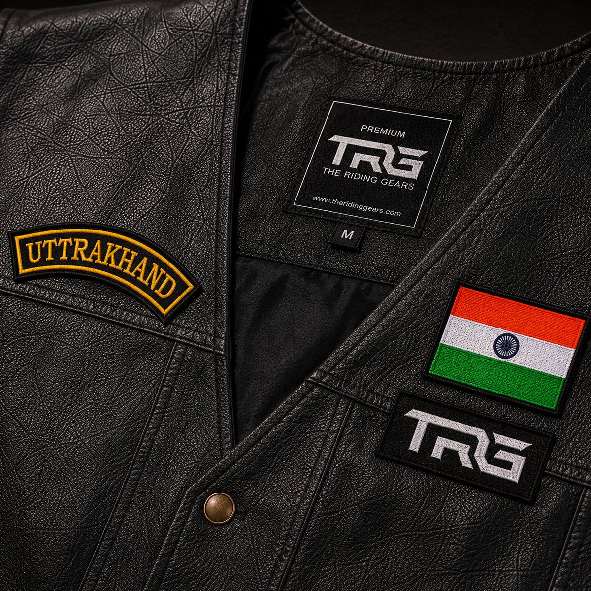 Close-up of Uttarakhand front curved embroidery patch with golden yellow lettering, black background and stitched border on a textured leather vest with Indian flag and TRG patches.